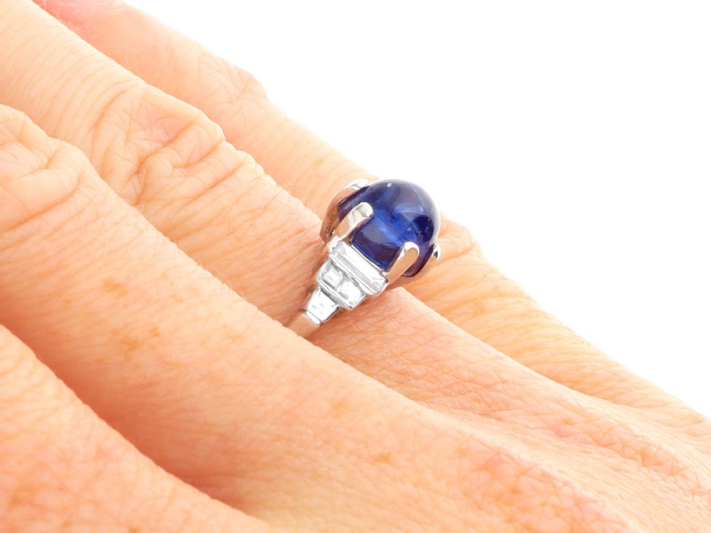 An Art Deco platinum dress ring set with a central oval cabochon blue sapphire flanked by stepped geometric shoulders of baguette cut diamonds. View XI