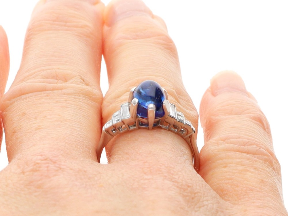 An Art Deco platinum dress ring set with a central oval cabochon blue sapphire flanked by stepped geometric shoulders of baguette cut diamonds. View XII