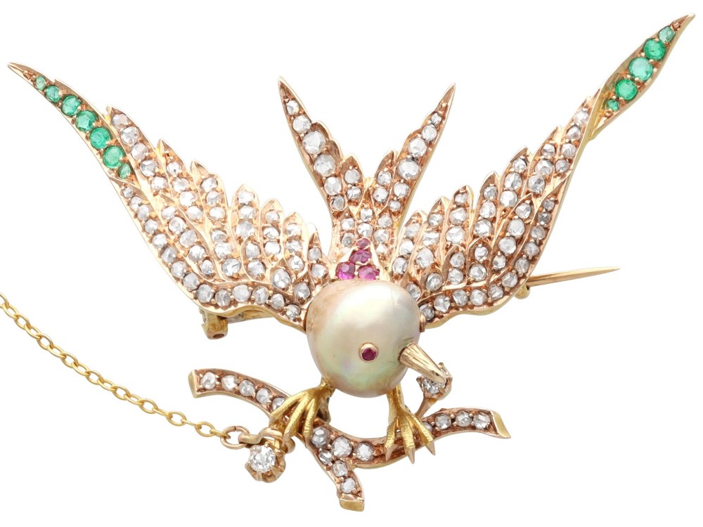 An antique Victorian 18ct yellow gold bird brooch set with diamonds on the wings, emeralds along the outer feathers, a natural pearl body, and ruby details.