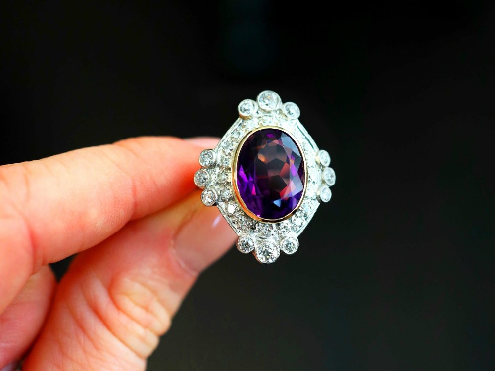An Art Deco ring featuring a central oval purple amethyst set in yellow gold, framed by a millegrain diamond halo. View V