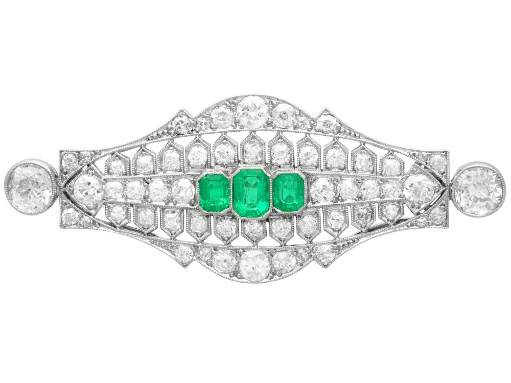 An Art Deco platinum brooch featuring three central step-cut green emeralds surrounded by a geometric lattice of diamonds with millegrain borders.