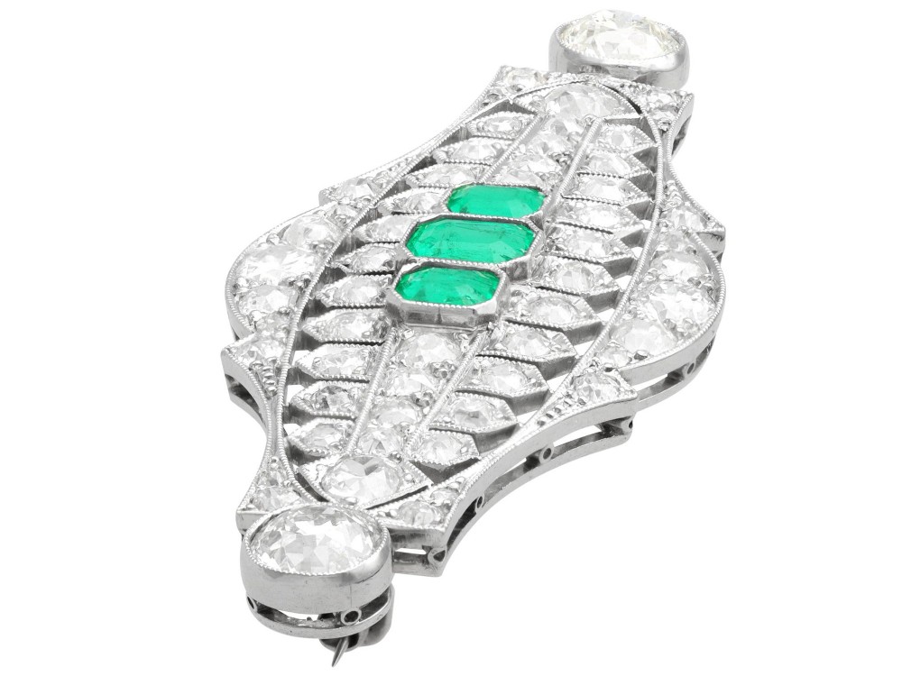 An Art Deco platinum brooch featuring three central step-cut green emeralds surrounded by a geometric lattice of diamonds with millegrain borders. View V