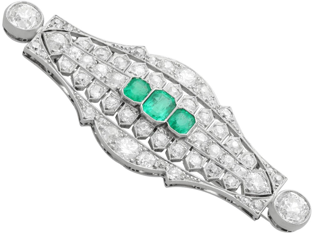 An Art Deco platinum brooch featuring three central step-cut green emeralds surrounded by a geometric lattice of diamonds with millegrain borders. View VI