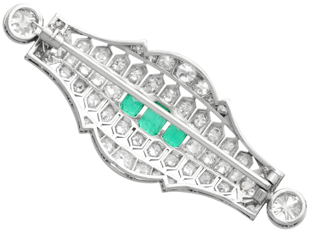 An Art Deco platinum brooch featuring three central step-cut green emeralds surrounded by a geometric lattice of diamonds with millegrain borders. View VII