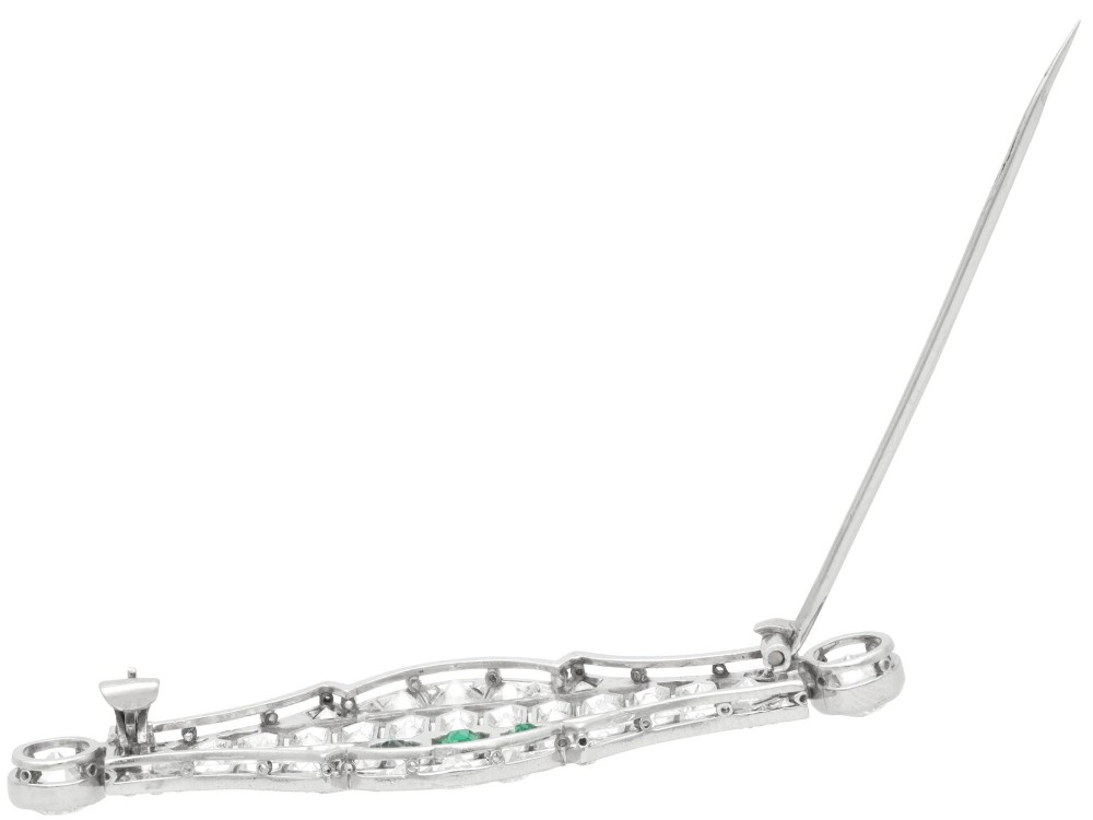 An Art Deco platinum brooch featuring three central step-cut green emeralds surrounded by a geometric lattice of diamonds with millegrain borders. View VIII