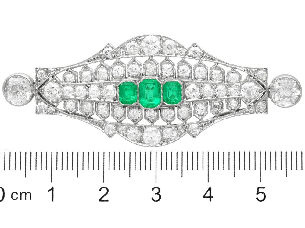 An Art Deco platinum brooch featuring three central step-cut green emeralds surrounded by a geometric lattice of diamonds with millegrain borders. View IX