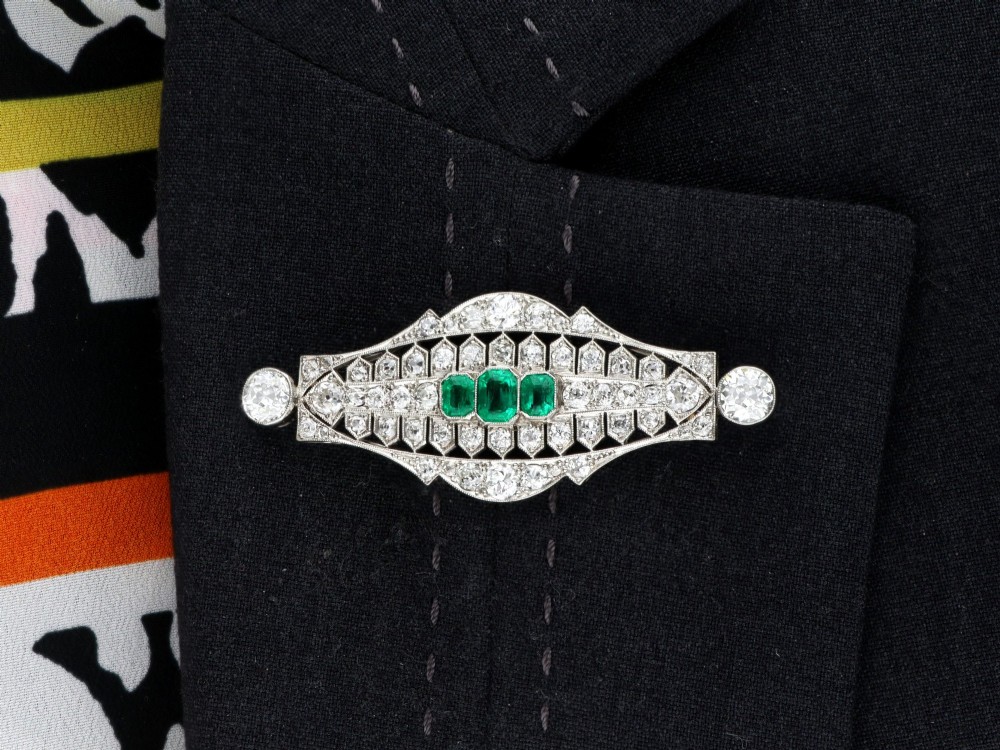 An Art Deco platinum brooch featuring three central step-cut green emeralds surrounded by a geometric lattice of diamonds with millegrain borders. View X