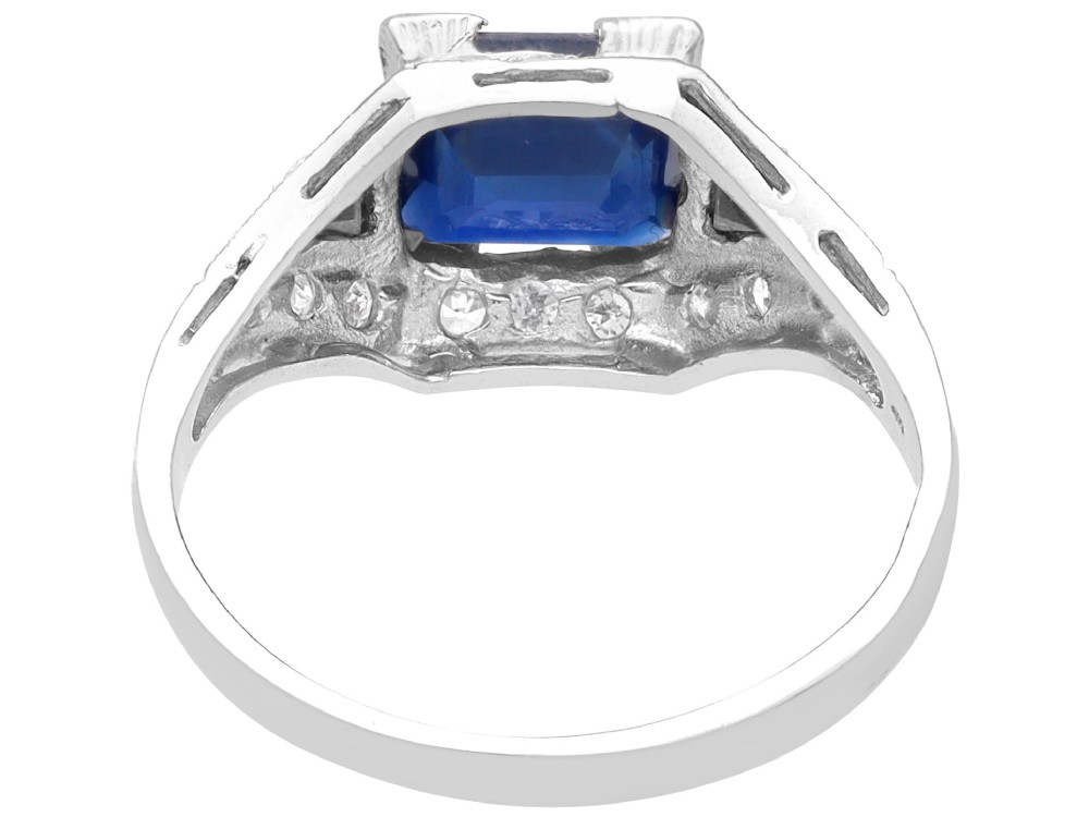 An Art Deco platinum ring featuring a central rectangular step-cut blue sapphire flanked by triangular sapphires and diamond-set shoulder details. View VI
