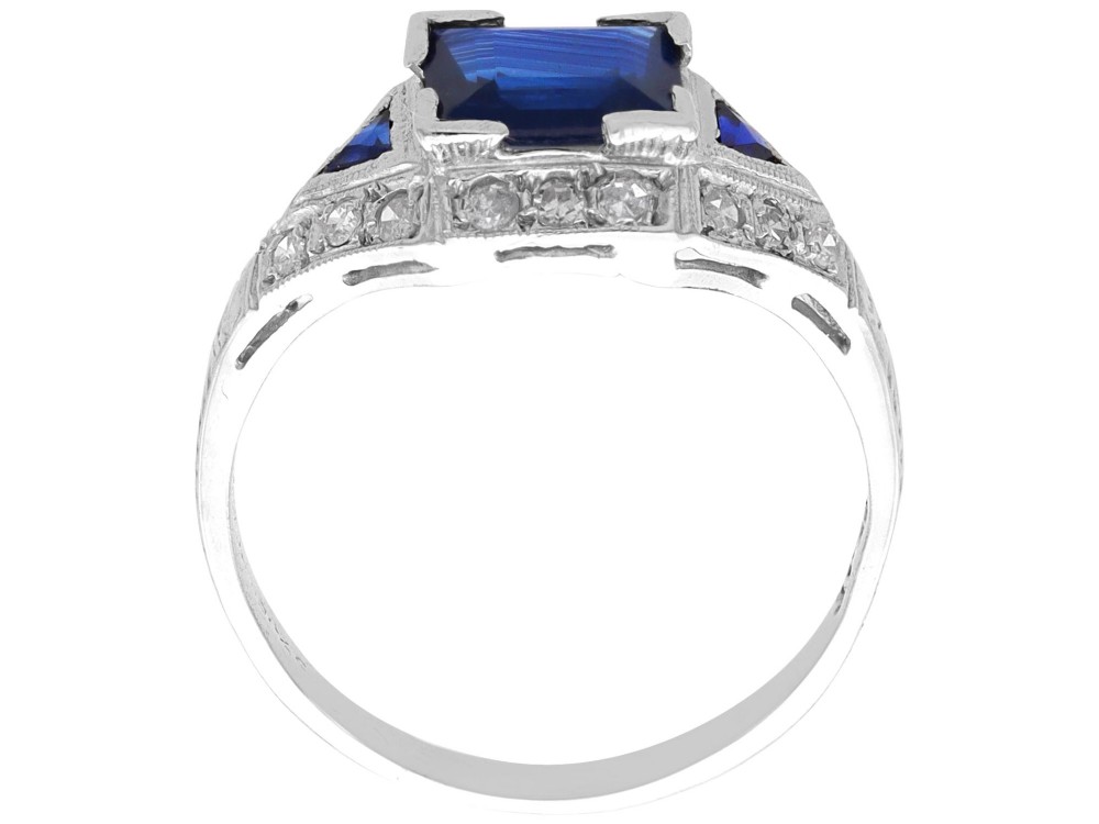 An Art Deco platinum ring featuring a central rectangular step-cut blue sapphire flanked by triangular sapphires and diamond-set shoulder details. View VII