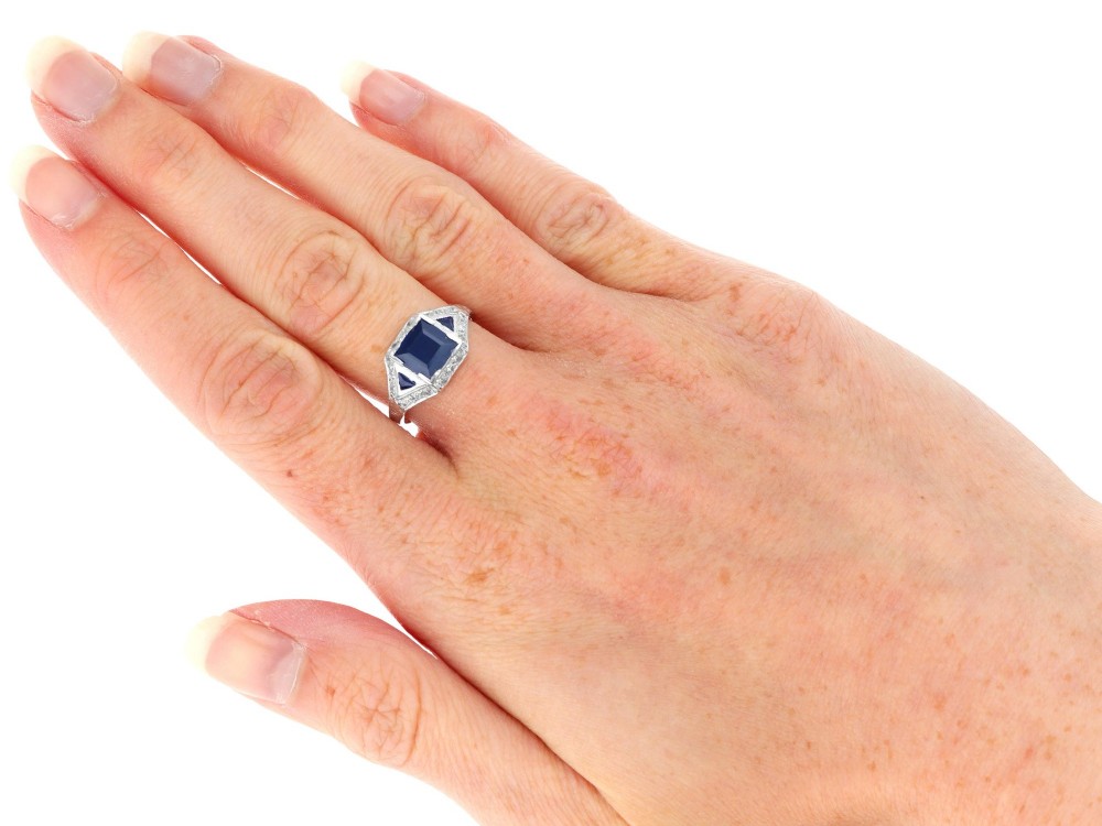 An Art Deco platinum ring featuring a central rectangular step-cut blue sapphire flanked by triangular sapphires and diamond-set shoulder details. View X