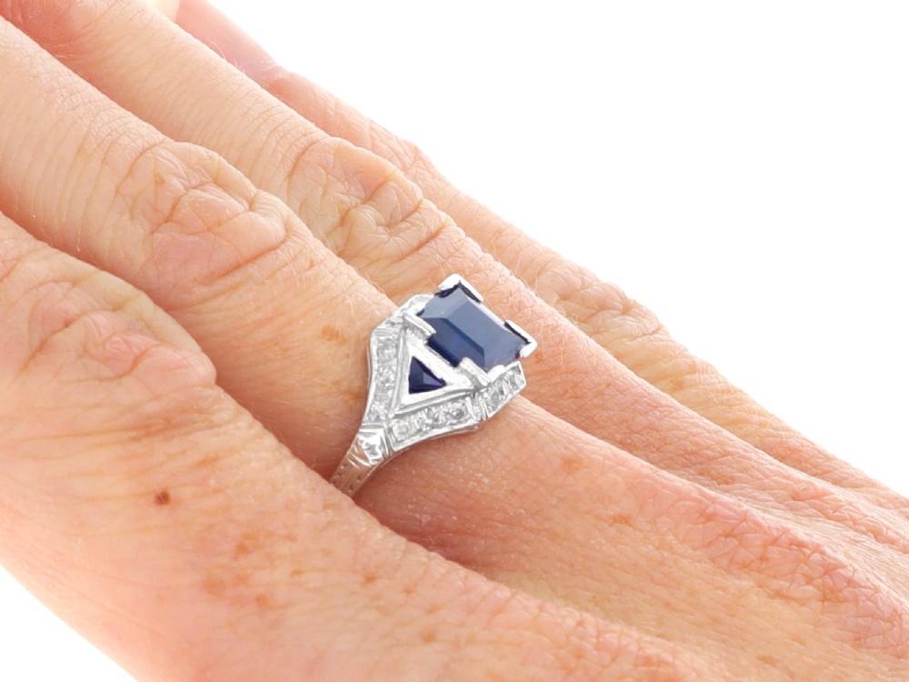 An Art Deco platinum ring featuring a central rectangular step-cut blue sapphire flanked by triangular sapphires and diamond-set shoulder details. View XI