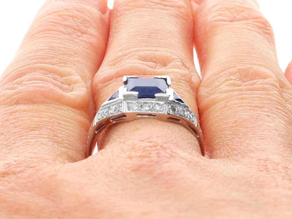 An Art Deco platinum ring featuring a central rectangular step-cut blue sapphire flanked by triangular sapphires and diamond-set shoulder details. View XII