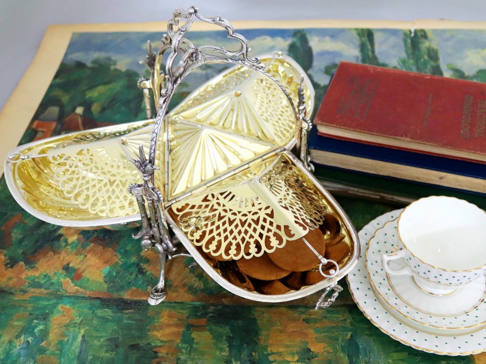 An antique Victorian sterling silver folding biscuit box featuring shell-form compartments, an ornate looped handle, and an engraved 1897 monogram. View VI