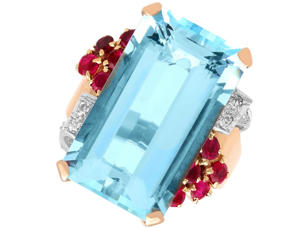 A 1940s Retro rose gold cocktail ring set with a large emerald-cut aquamarine flanked by ruby clusters and diamond accents.