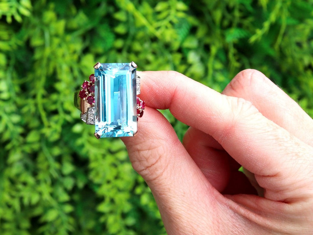 A 1940s Retro rose gold cocktail ring set with a large emerald-cut aquamarine flanked by ruby clusters and diamond accents. View II