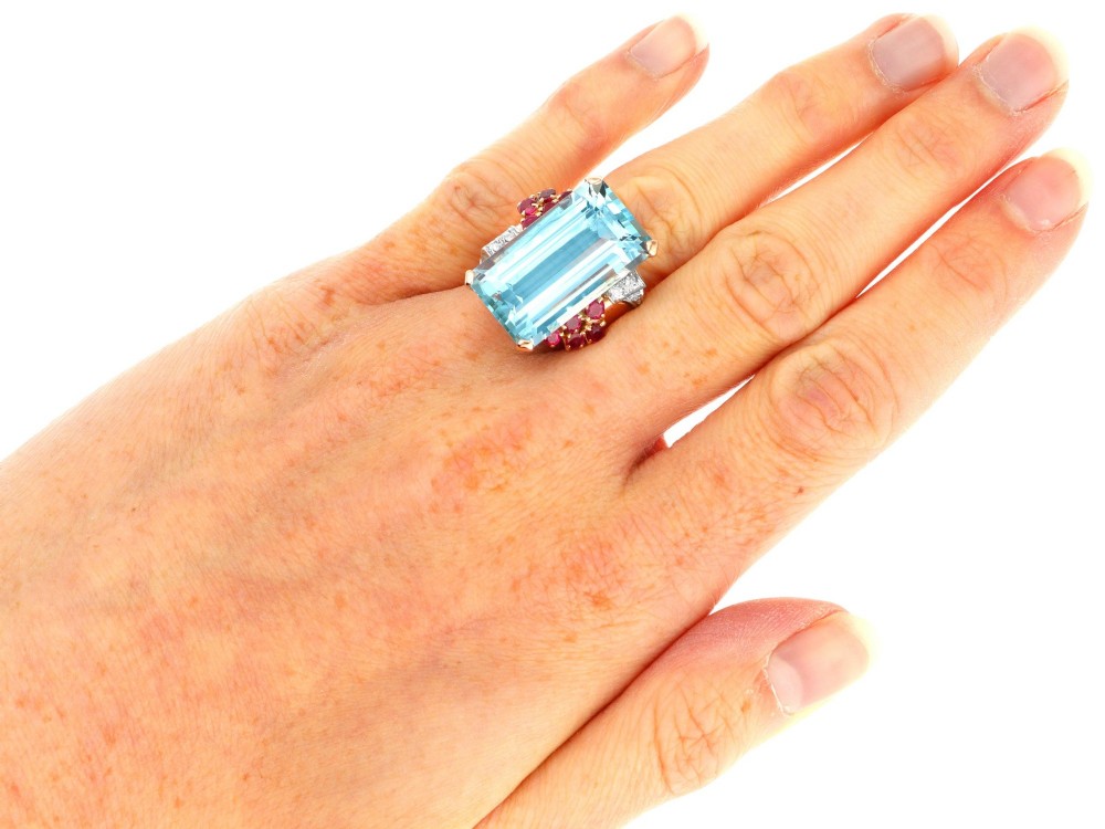 A 1940s Retro rose gold cocktail ring set with a large emerald-cut aquamarine flanked by ruby clusters and diamond accents. View X