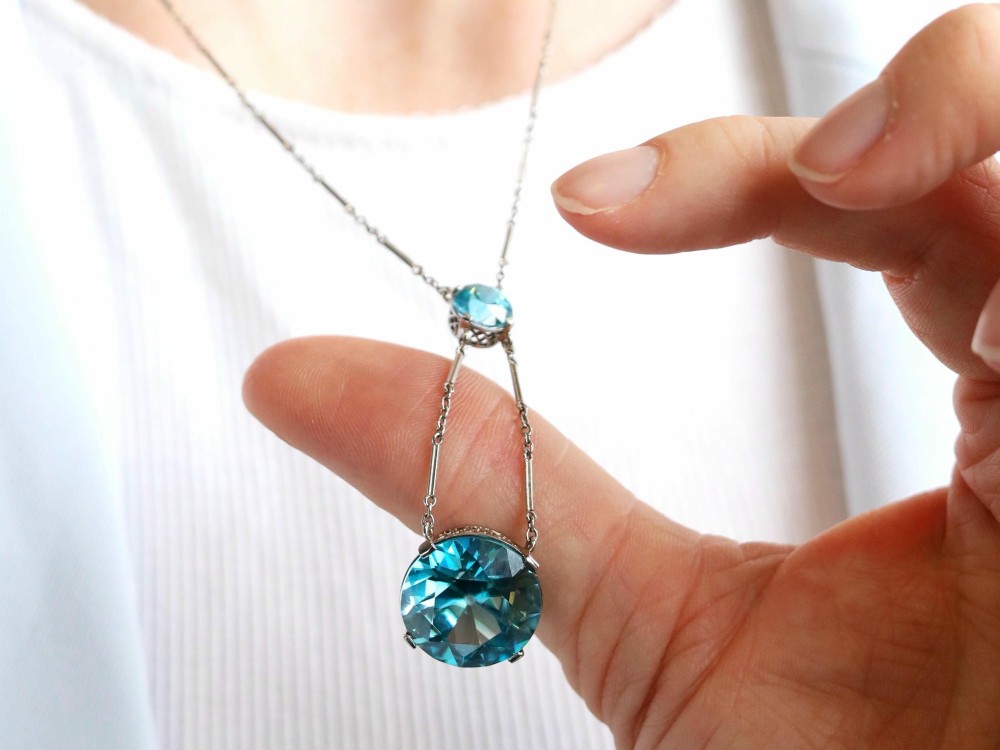 An Art Deco 18ct white gold drop pendant set with two round brilliant-cut bright blue zircon gemstones suspended on delicate bar links. View V
