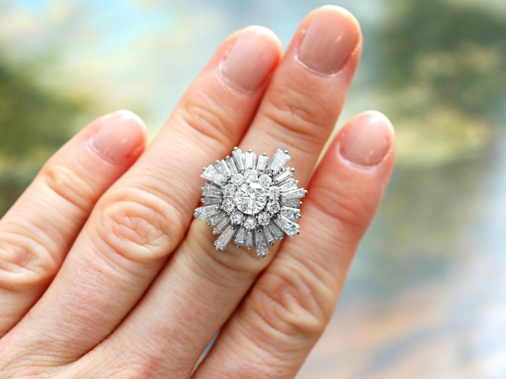 A vintage 18ct white gold cluster ring featuring a central round brilliant cut diamond surrounded by a halo of brilliant cut diamonds and an outer sunburst tier of tapered baguette diamonds. View IV