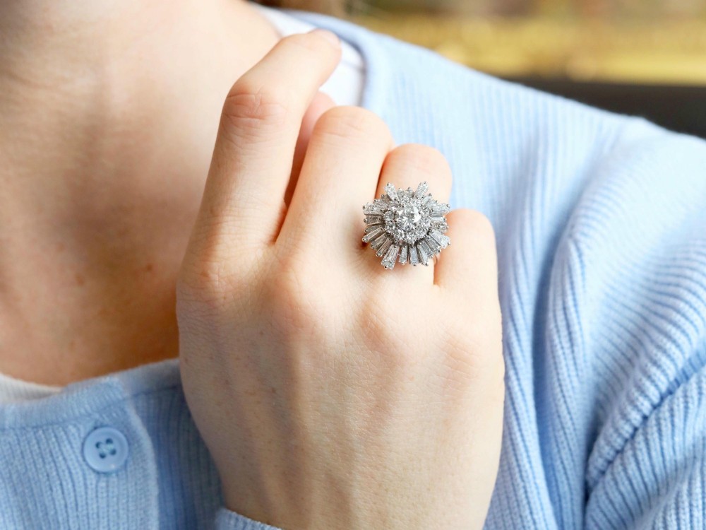 A vintage 18ct white gold cluster ring featuring a central round brilliant cut diamond surrounded by a halo of brilliant cut diamonds and an outer sunburst tier of tapered baguette diamonds. View V