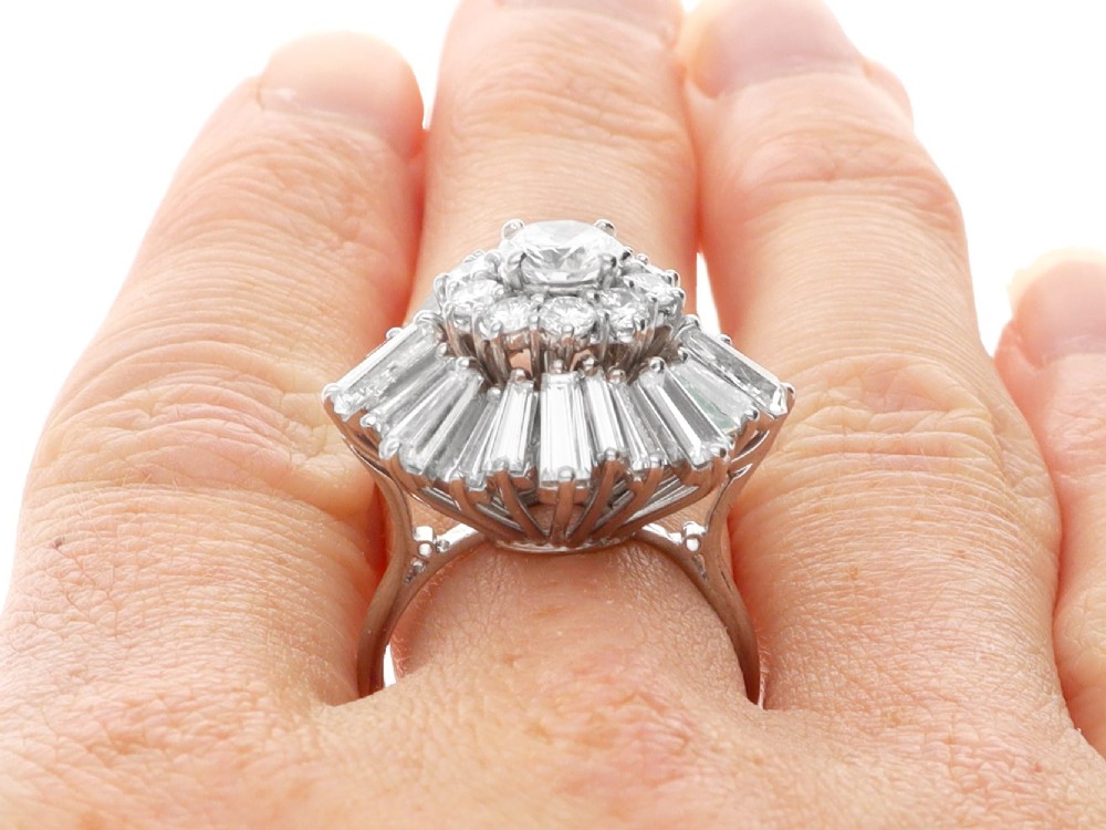 A vintage 18ct white gold cluster ring featuring a central round brilliant cut diamond surrounded by a halo of brilliant cut diamonds and an outer sunburst tier of tapered baguette diamonds. View XII