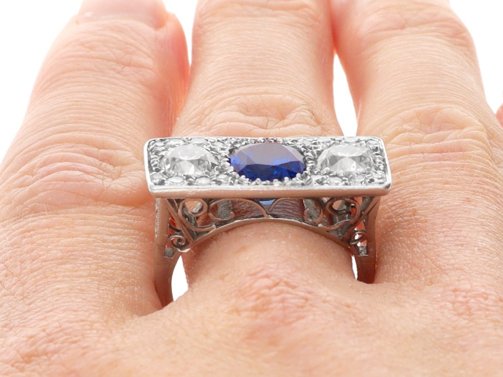 An Art Deco platinum plaque ring set with a central blue sapphire flanked by two large diamonds and surrounded by smaller pave-set diamonds. View X