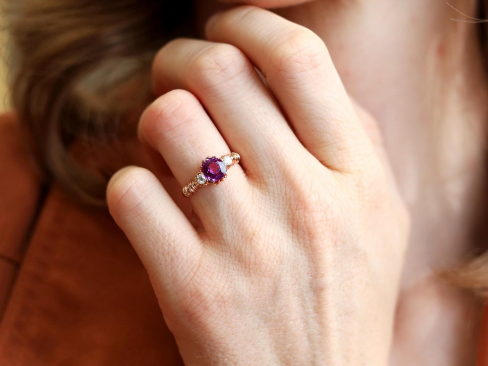 A Victorian 15ct rose gold trilogy ring set with a central purple sapphire flanked by two diamonds on carved shoulders. View IV