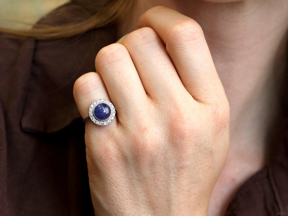An Art Deco platinum cluster ring featuring a central cabochon star sapphire encircled by a halo of round diamonds with millegrain edging. View IV