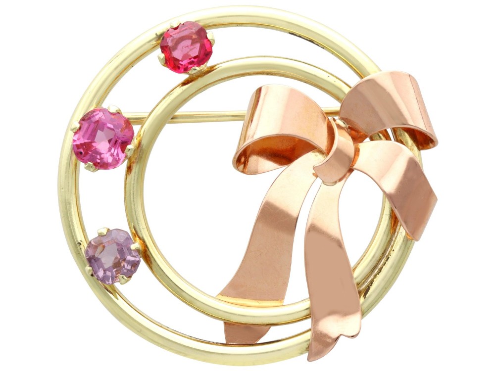 A circular two-tone gold brooch featuring yellow gold concentric rings set with three pink spinels and decorated with a rose gold bow.
