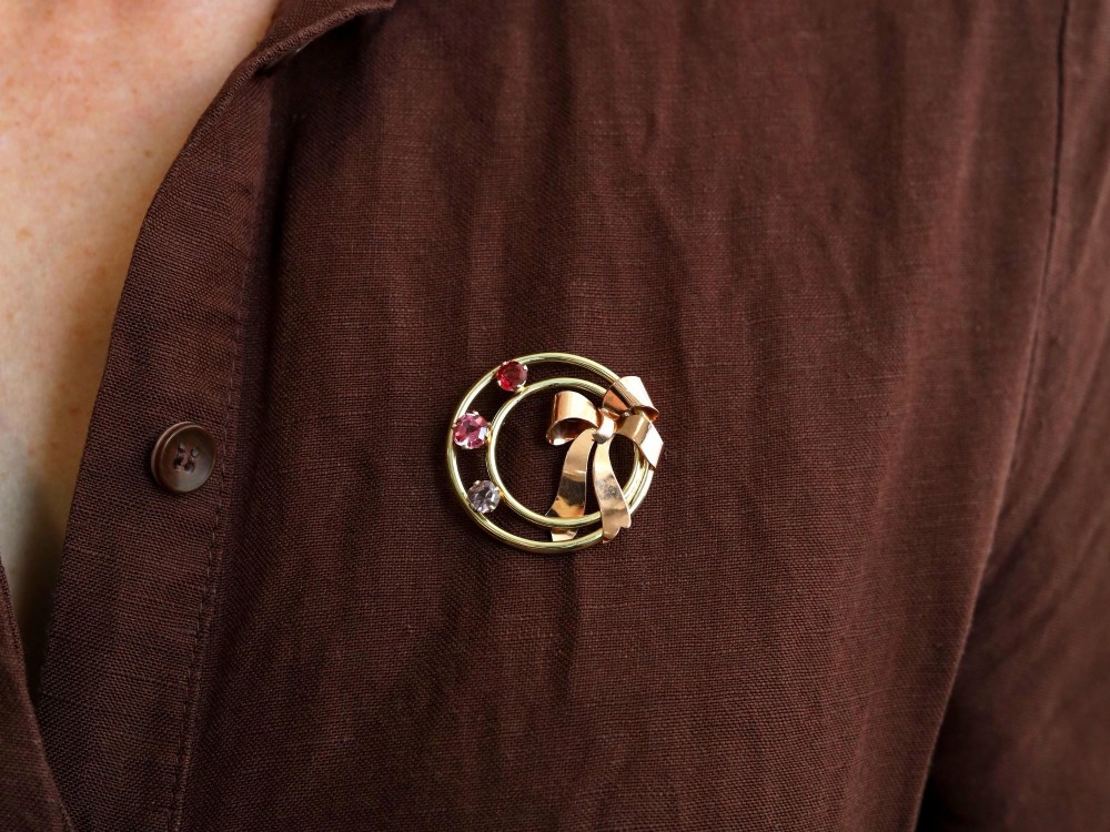 A circular two-tone gold brooch featuring yellow gold concentric rings set with three pink spinels and decorated with a rose gold bow. View V