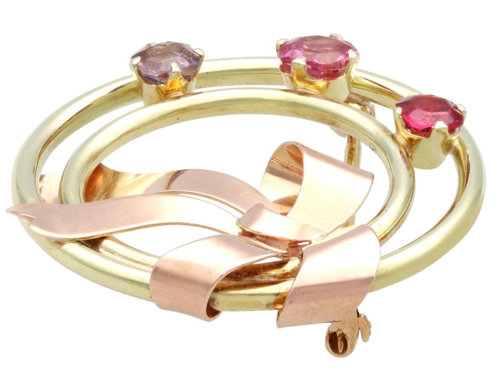 A circular two-tone gold brooch featuring yellow gold concentric rings set with three pink spinels and decorated with a rose gold bow. View IX