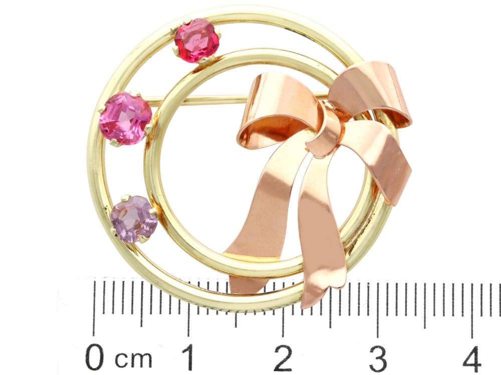 A circular two-tone gold brooch featuring yellow gold concentric rings set with three pink spinels and decorated with a rose gold bow. View XIII