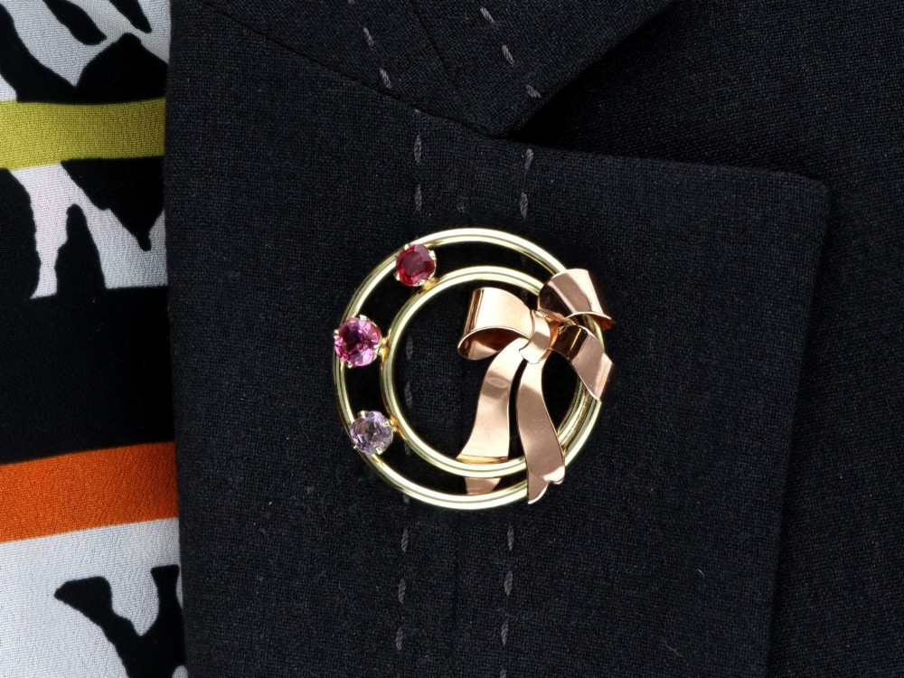 A circular two-tone gold brooch featuring yellow gold concentric rings set with three pink spinels and decorated with a rose gold bow. View XV
