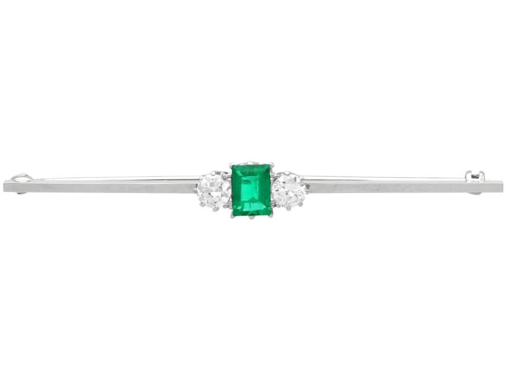 An antique 18ct white gold bar brooch featuring a central rectangular emerald cut emerald flanked by two round diamonds on a slender horizontal metal bar.
