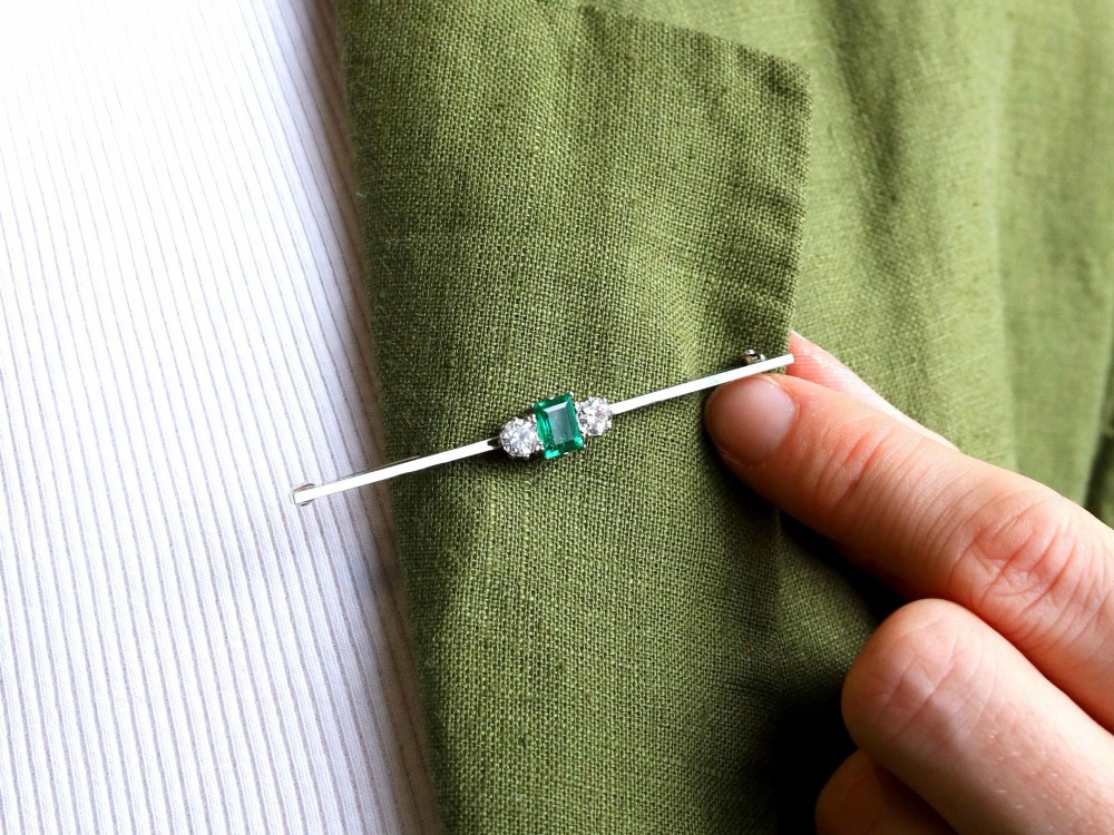 An antique 18ct white gold bar brooch featuring a central rectangular emerald cut emerald flanked by two round diamonds on a slender horizontal metal bar. View IV