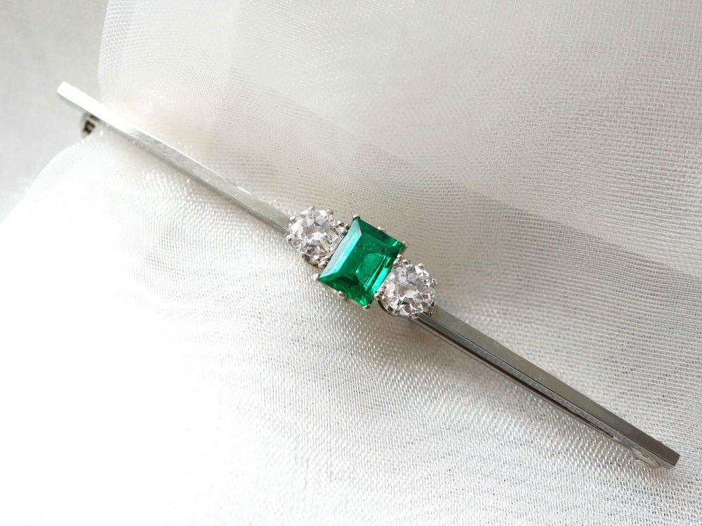 An antique 18ct white gold bar brooch featuring a central rectangular emerald cut emerald flanked by two round diamonds on a slender horizontal metal bar. View V