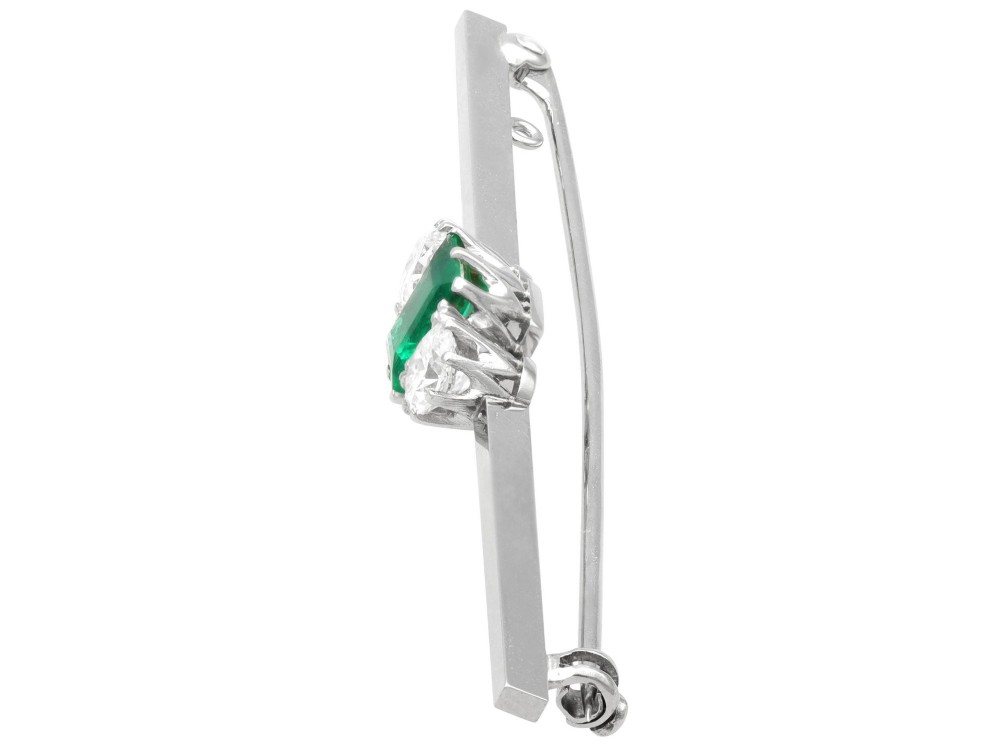 An antique 18ct white gold bar brooch featuring a central rectangular emerald cut emerald flanked by two round diamonds on a slender horizontal metal bar. View VII