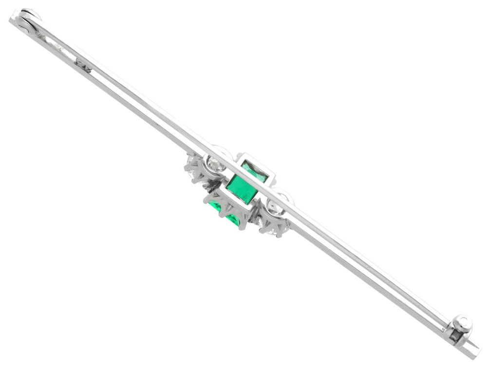 An antique 18ct white gold bar brooch featuring a central rectangular emerald cut emerald flanked by two round diamonds on a slender horizontal metal bar. View VIII