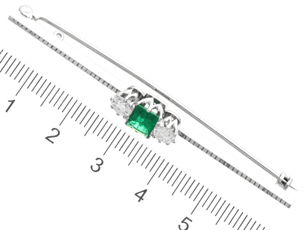 An antique 18ct white gold bar brooch featuring a central rectangular emerald cut emerald flanked by two round diamonds on a slender horizontal metal bar. View X