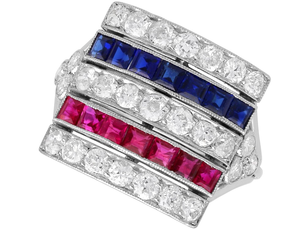 An antique platinum ring displaying horizontal rows of round diamonds alternating with channel-set rectangular blue sapphires and red rubies.
