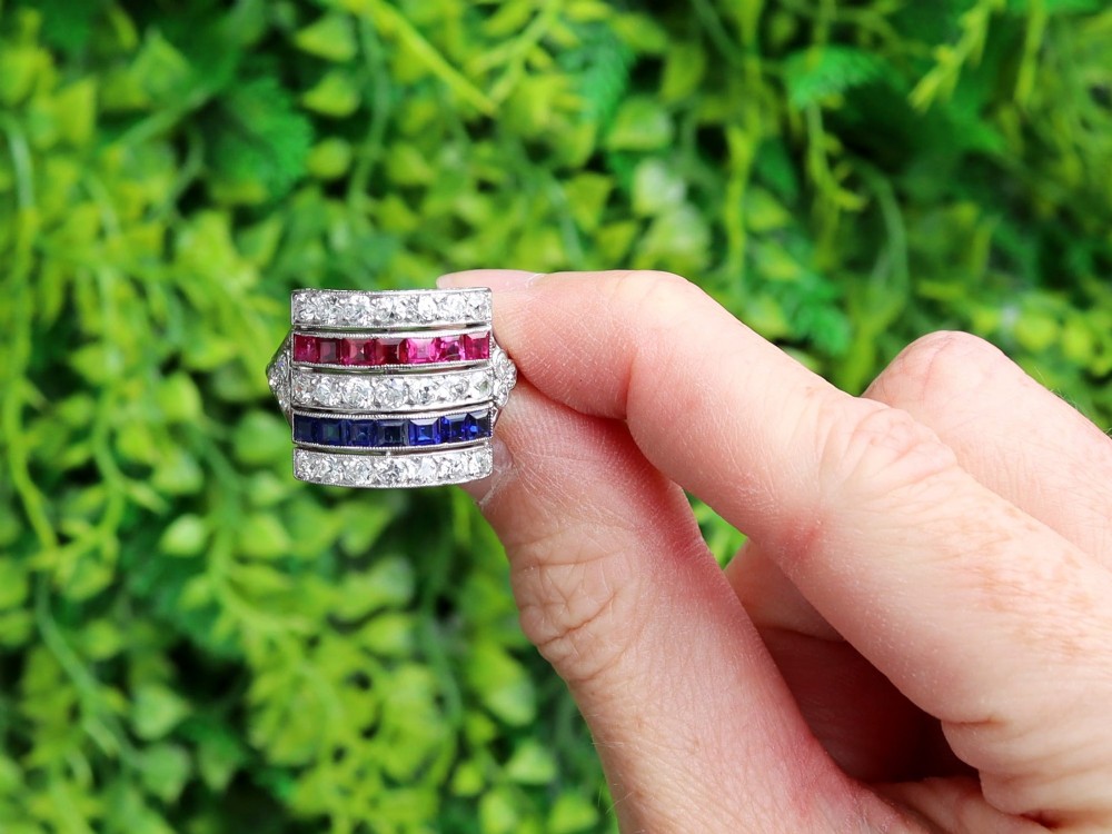 An antique platinum ring displaying horizontal rows of round diamonds alternating with channel-set rectangular blue sapphires and red rubies. View II