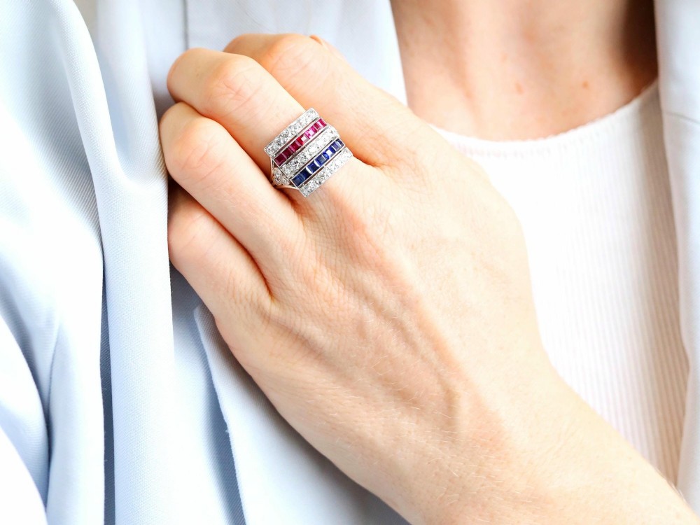 An antique platinum ring displaying horizontal rows of round diamonds alternating with channel-set rectangular blue sapphires and red rubies. View IV