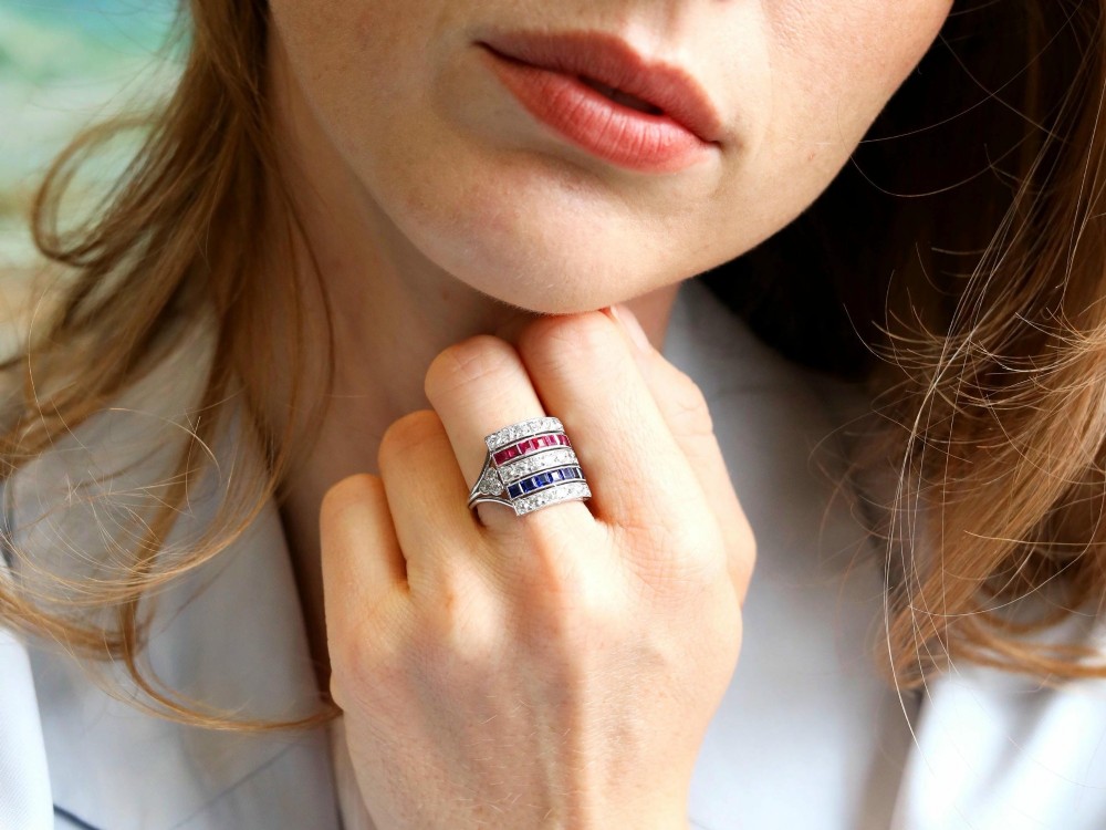 An antique platinum ring displaying horizontal rows of round diamonds alternating with channel-set rectangular blue sapphires and red rubies. View V