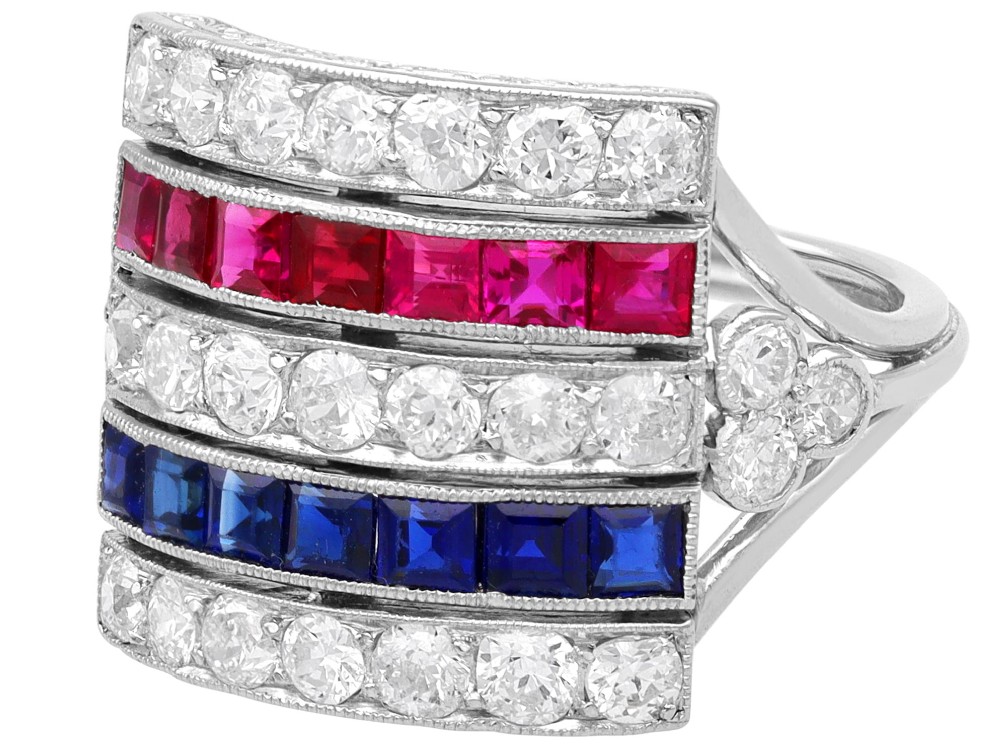 An antique platinum ring displaying horizontal rows of round diamonds alternating with channel-set rectangular blue sapphires and red rubies. View VII