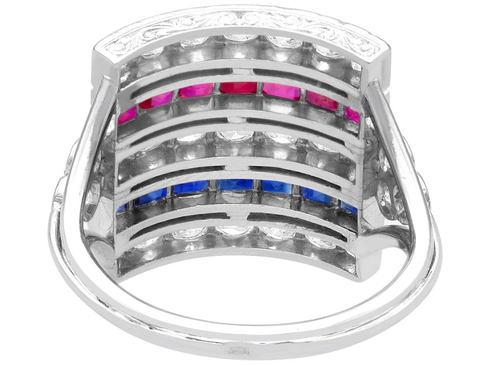 An antique platinum ring displaying horizontal rows of round diamonds alternating with channel-set rectangular blue sapphires and red rubies. View VIII