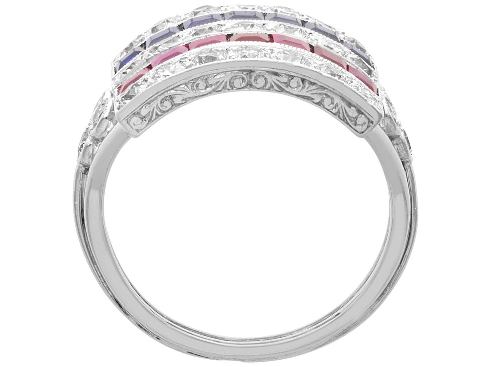 An antique platinum ring displaying horizontal rows of round diamonds alternating with channel-set rectangular blue sapphires and red rubies. View IX