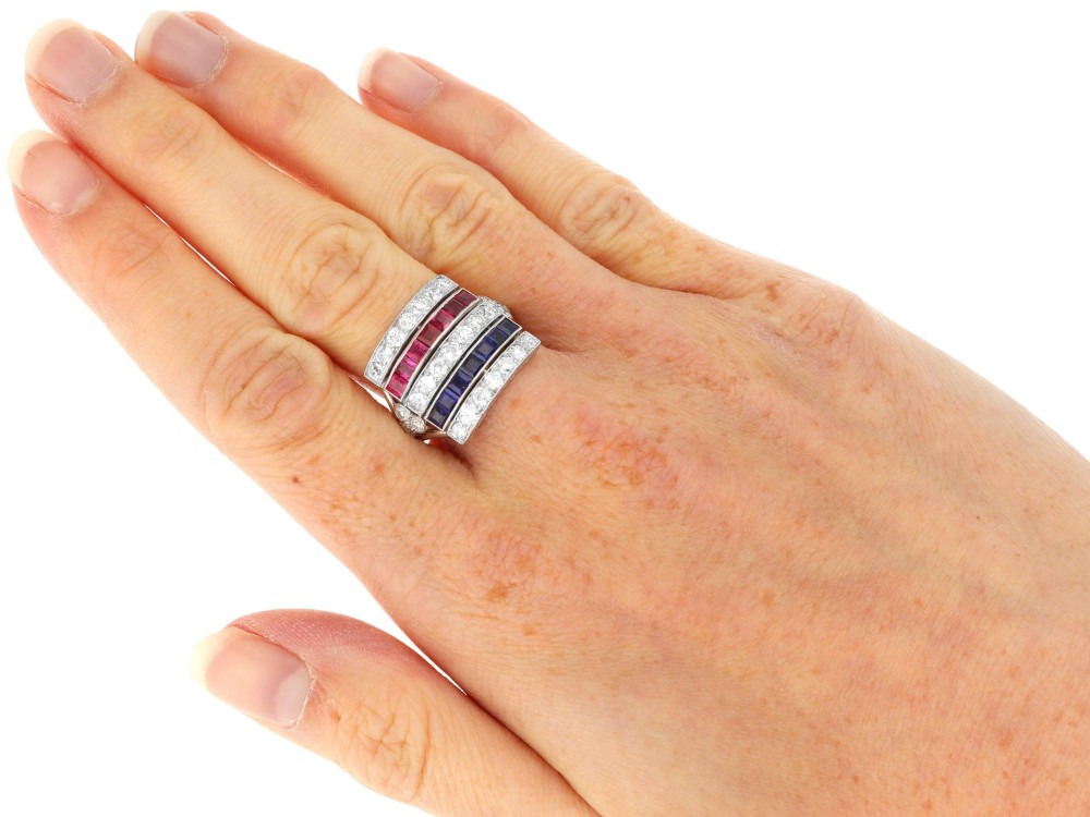 An antique platinum ring displaying horizontal rows of round diamonds alternating with channel-set rectangular blue sapphires and red rubies. View XI
