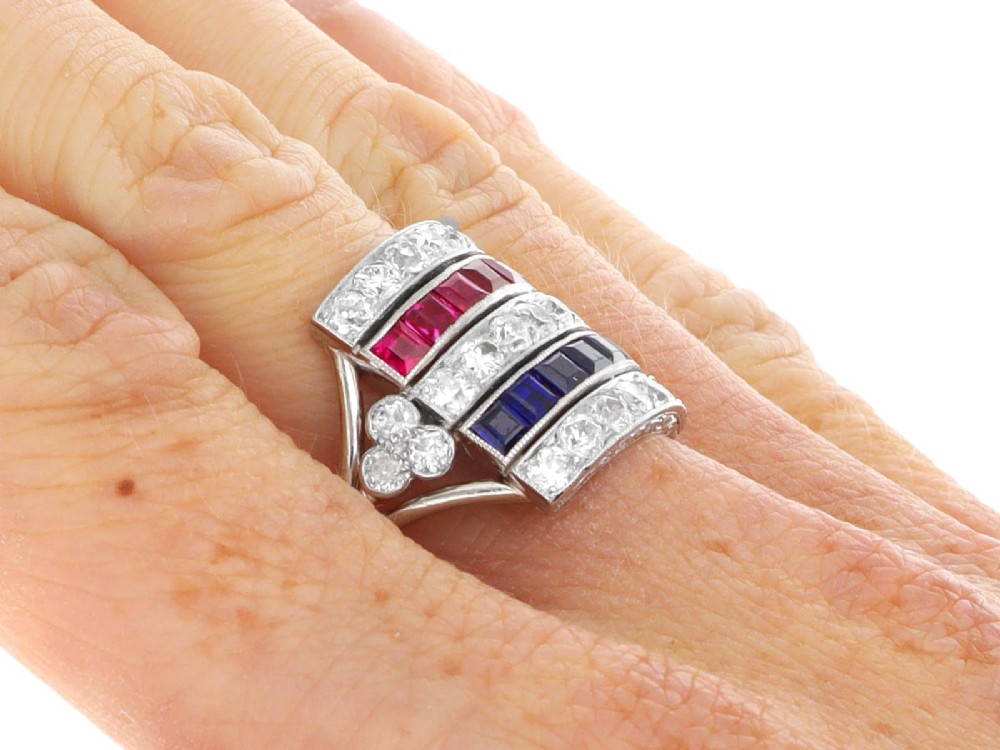 An antique platinum ring displaying horizontal rows of round diamonds alternating with channel-set rectangular blue sapphires and red rubies. View XII