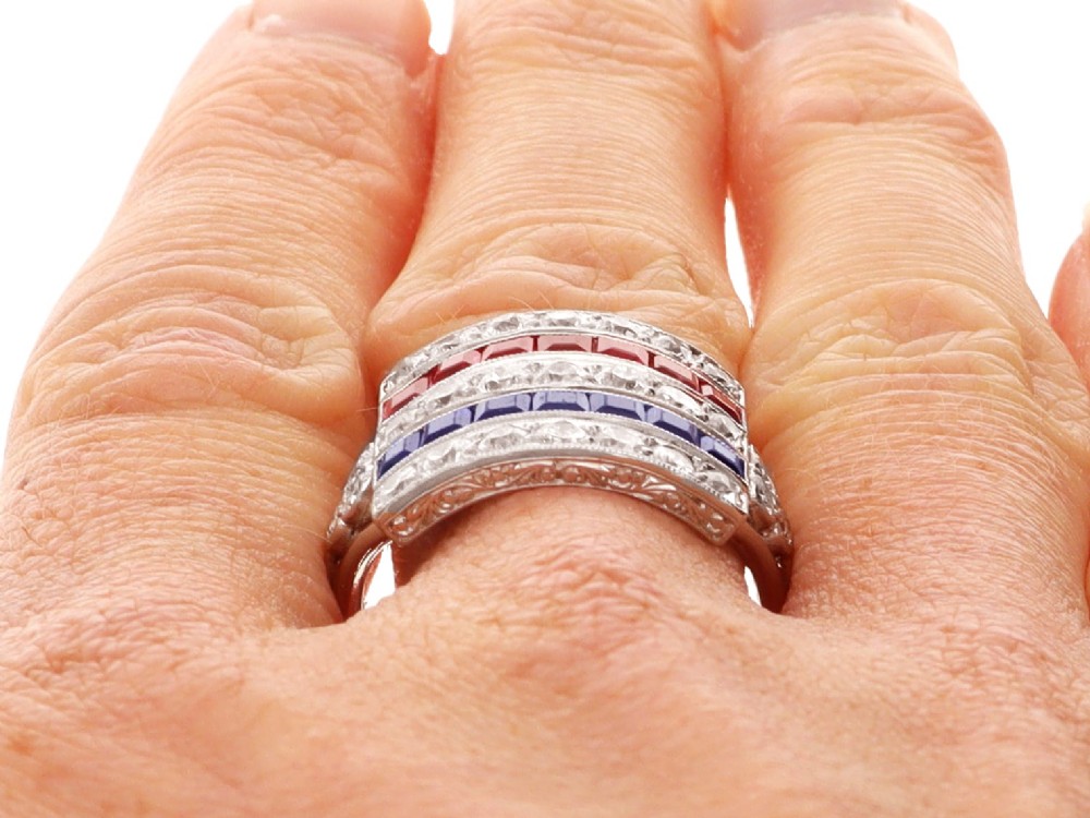 An antique platinum ring displaying horizontal rows of round diamonds alternating with channel-set rectangular blue sapphires and red rubies. View XIII
