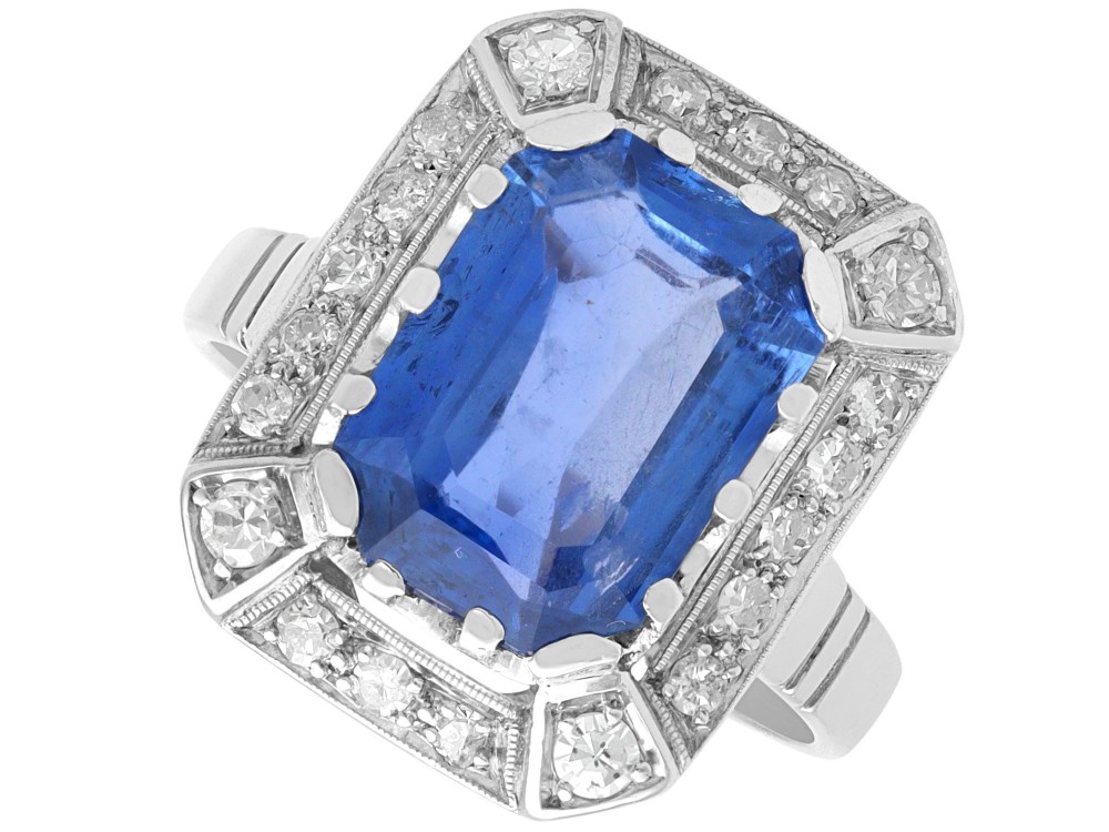 An Art Deco 18ct white gold ring featuring a central octagonal step-cut blue Ceylon sapphire surrounded by a geometric diamond halo with milgrain details.