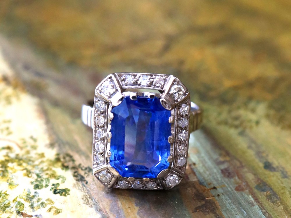 An Art Deco 18ct white gold ring featuring a central octagonal step-cut blue Ceylon sapphire surrounded by a geometric diamond halo with milgrain details. View V